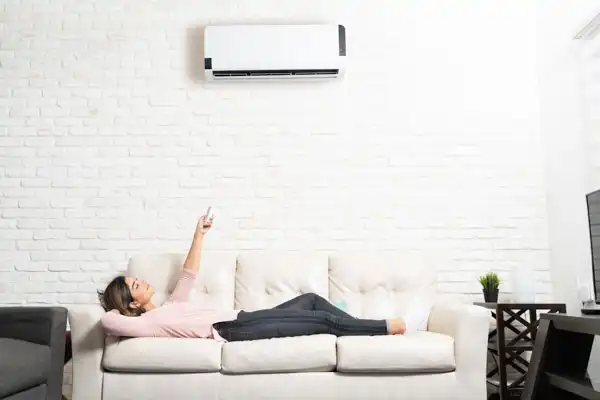 A woman lies on a white sofa and points a remote control at a wall-mounted air conditioner, enjoying the comfort provided by recent mini-split installation in her bright living room.