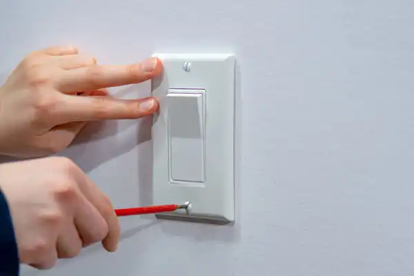 A person in Goshen NY uses a screwdriver to unscrew the bottom of a white electrical switch cover plate, with one hand steadying the plate—demonstrating professional outlets service.