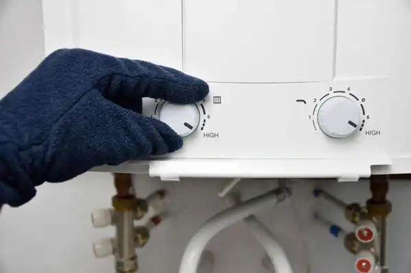 A gloved hand adjusts the temperature dial to high on a white appliance—likely during tankless water heater maintenance—in a Goshen, NY home, with pipes and hoses visible underneath.