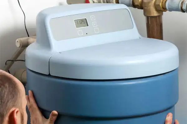 A person performs a water softener installation in Goshen, NY, adjusting a blue tank with a digital control panel in a utility area.