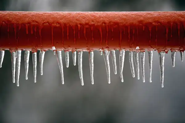 Close-up of a red metal pipe with numerous icicles hanging from its underside, highlighting the need for frozen pipe repair in Goshen NY, against a blurred background.