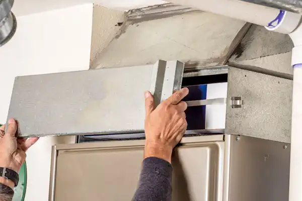 A person performs furnace maintenance in Goshen NY by removing the metal cover from a furnace to access and replace the air filter inside.