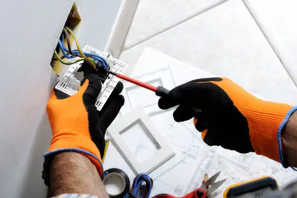 A person in orange gloves uses a screwdriver to perform electrical repair on an outlet in Goshen, NY, with tools and outlet covers nearby against a white wall.