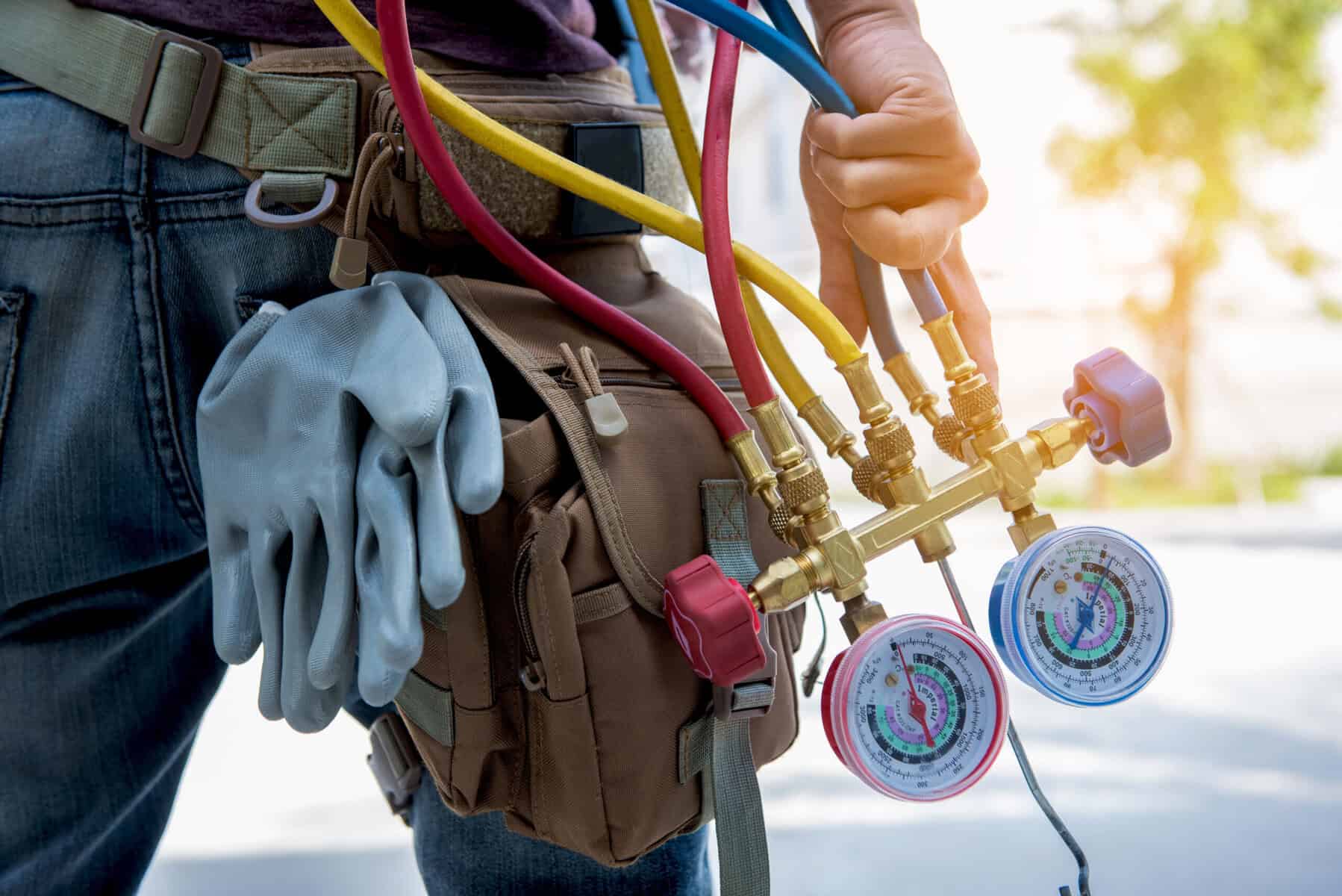 A person holding HVAC manifold gauges and wearing a tool bag with work gloves attached, standing outdoors—ready to provide top-notch AC services in Goshen NY.