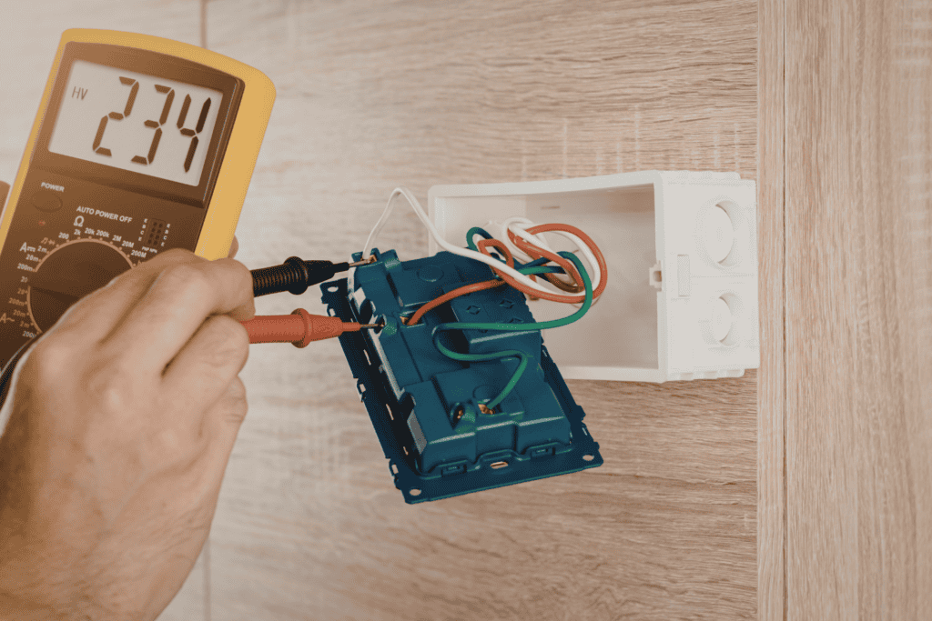 A person uses a digital multimeter to measure voltage on exposed wiring inside a wall-mounted electrical box during a safety inspection.